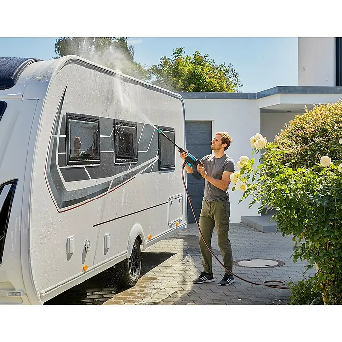 Gardena Power for All 18V Akku-Druckreiniger AquaClean 24/18V SE - 18 V, 1 Akku, 2,5 Ah - Ein Mann reinigt mit einem Gardena Akku-Hochdruckreiniger eine Wohnwagenwand, umgeben von Grün und einem modernen Haus im Hintergrund.