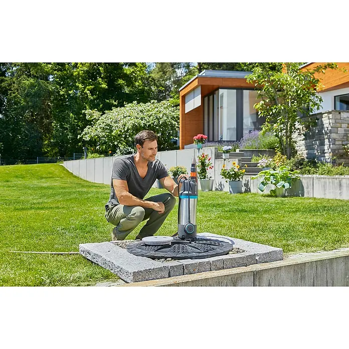 Gardena Bomba sumergible de presión 5900/4 Inox - Capacidad de bombeo máx.: 5.900 l/h, Altura de transporte máx.: 35 m - Un hombre está en cuclillas junto a una bomba de jardín Gardena plateada en un pozo pavimentado sobre un césped verde. Hermosa casa moderna al fondo.