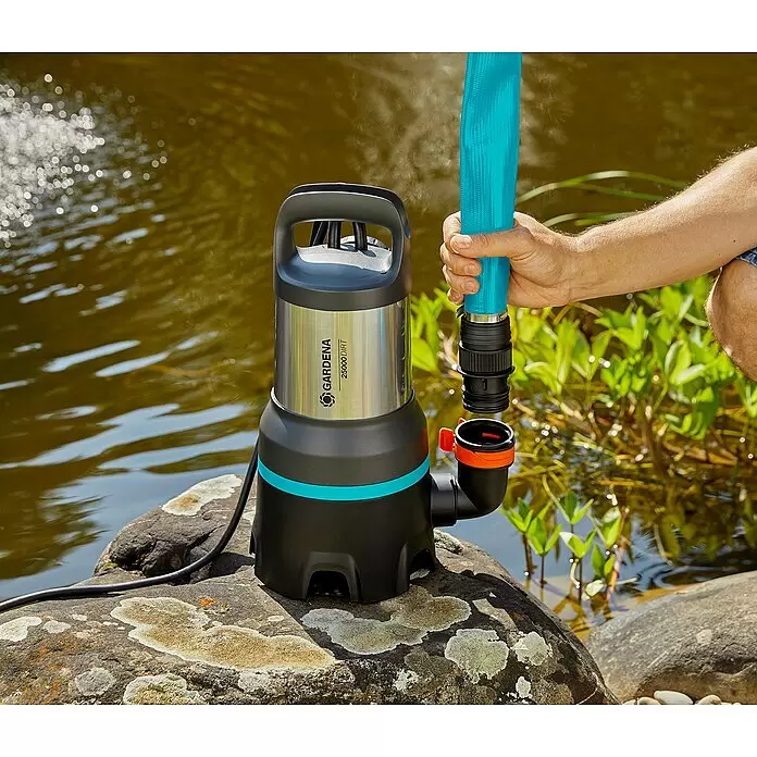 Gardena Schmutzwasserpumpe 25000 - 1.100 W, Max. Fördermenge: 25.000 l/h, Eintauchtiefe: 7 m - Eine Person schließt einen blauen Schlauch an eine schwarze und Edelstahl GARDENA 25000 DIRT Pumpe auf einem Felsen neben Wasser an.