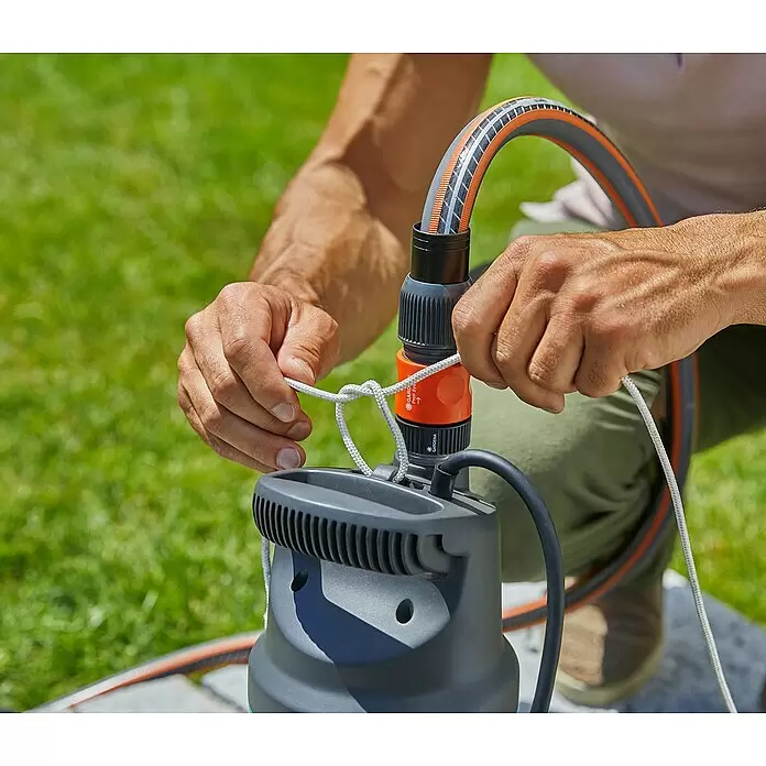 Gardena Tauchpumpe 5900/4 INOX Automatic - 900 W, Max. Fördermenge: 5.900 l/h - Hände verbinden einen grau-orangen Gartenschlauch mit einer grauen Tauchpumpe und befestigen sie mit einem weißen Seil. Die Pumpe wird für den Außeneinsatz vorbereitet.