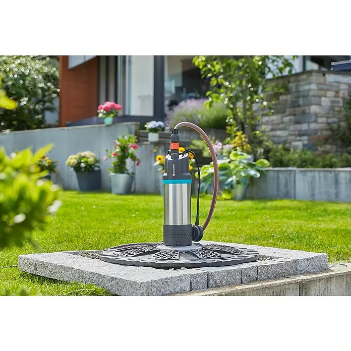 Gardena Drucktauchpumpe 5900/4 Inox - 900 W, Max. Fördermenge: 5.900 l/h - Eine Gardena Tauchdruckpumpe steht auf einer Steinfläche inmitten eines üppigen Gartens, angeschlossen an einen orangefarbenen Schlauch.