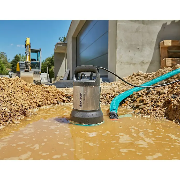 Gardena Schmutzwasserpumpe 25000 - 1.100 W, Max. Fördermenge: 25.000 l/h, Eintauchtiefe: 7 m - Die Gardena Schmutzwasser-Tauchpumpe 25000 Dirt steht im trüben Wasser einer Baustelle, angeschlossen an einen blauen Schlauch.