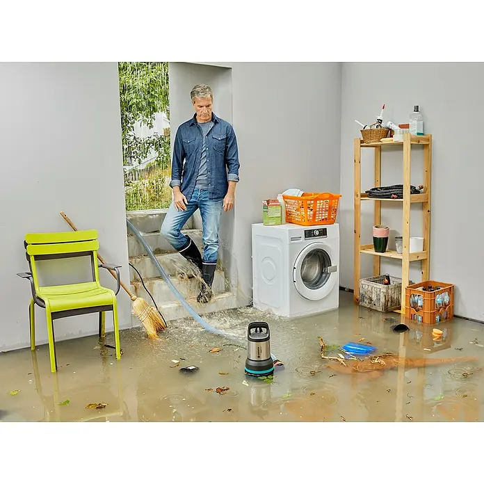 Gardena Schmutzwasserpumpe 25000 - 1.100 W, Max. Fördermenge: 25.000 l/h, Eintauchtiefe: 7 m - Ein Mann steht in einem überfluteten Raum mit einer GARDENA-Tauchpumpe, die Wasser ableitet. Die Pumpe ist blau und grau.