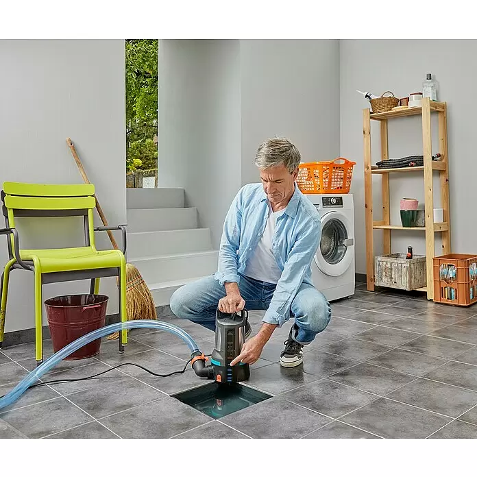 Gardena Klarwasser-Tauchpumpe 17000 Aquasensor - 750 W, Max. Fördermenge: 17.000 l/h - Mann hockt und bedient eine dunkelgraue und blaue Tauchpumpe mit durchsichtigem Schlauch, um Wasser aus einem Bodeneinlass abzupumpen.