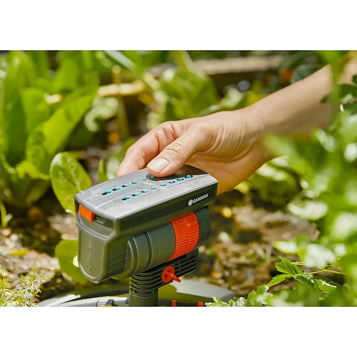 Gardena Viereckregner KopfPassend für: Gardena Viereckregner OS 90 und OS 140 - Eine Hand bedient einen Gardena-Regner, der im Garten steht. Das Gerät ist grau mit orangen Akzenten.