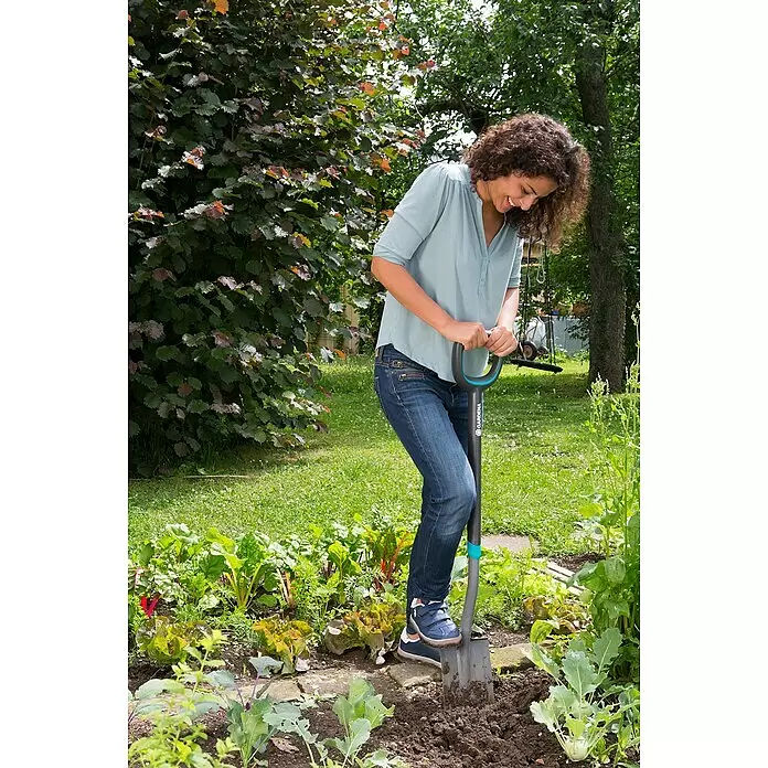 Gardena Ergoline Gärtnerspaten Damen - Mit Tritt, Arbeitsbreite: 16,4 cm - Eine Frau gräbt mit einer dunkelgrau-blauen Schaufel in einem üppigen Gartenbeet.