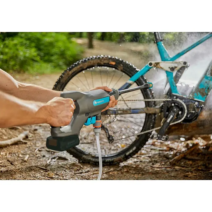 Gardena Power for All 18V Akku-Druckreiniger AquaClean 24/18V PR - 18 V, 1 Akku, 2,5 Ah - Ein Mann reinigt ein schlammiges Fahrrad mit einem tragbaren Gardena-Reiniger, der mit einer Batterie betrieben wird und einen Schlauchanschluss hat.