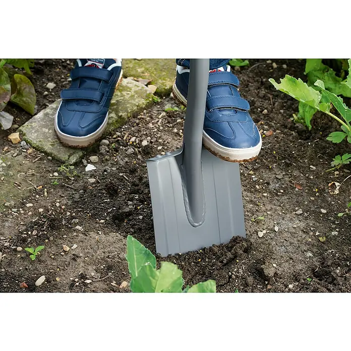 Gardena Ergoline Gärtnerspaten Damen - Mit Tritt, Arbeitsbreite: 16,4 cm - Nahaufnahme eines Spatens, der mit einem Fuß in einem Garten mit Erde, Pflanzen und Steinen in den Boden getreten wird.