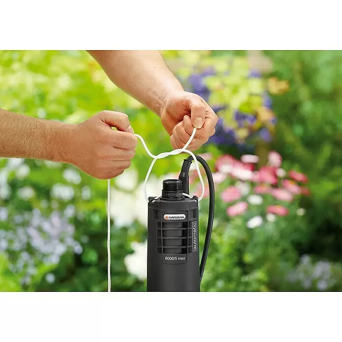 Gardena Tiefbrunnenpumpe 6000/5 Inox automatic - 950 W, Max. Fördermenge: 6.000 l/h, Max. Druck: 5 bar - Hände befestigen ein weißes Seil an einer schwarzen Gardena 6000/5 inox automatic Tauchpumpe, bereit für den Einsatz im Garten.