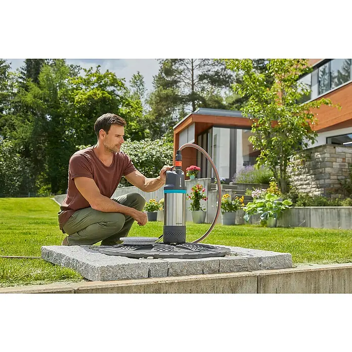 Gardena Tauchpumpe 5900/4 INOX Automatic - 900 W, Max. Fördermenge: 5.900 l/h - Ein Mann hockt neben einer silbernen GARDENA-Gartenpumpe und überprüft sie, im Hintergrund ein Garten mit üppigem Grün.