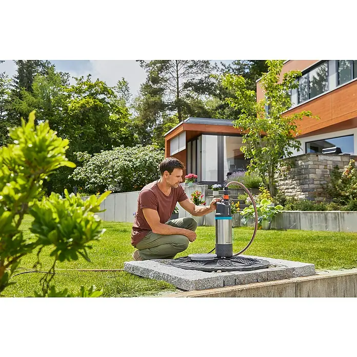 Gardena Tauchpumpe 5900/4 INOX Automatic - 900 W, Max. Fördermenge: 5.900 l/h - Ein Mann hockt neben einer Gardena-Tauchdruckpumpe auf einer Steinplattform, umgeben von üppigem Grün, und bedient sie.