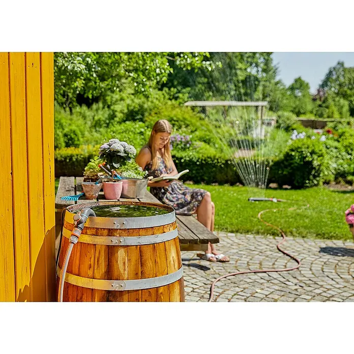 Gardena Regenfasspumpe 4700/2 INOX - 550 W, Max. Fördermenge: 4.700 l/h - Nahaufnahme eines Holzfasses zur Regenwassernutzung mit einem Schlauchanschluss in einem Garten, während eine Frau im Hintergrund liest.