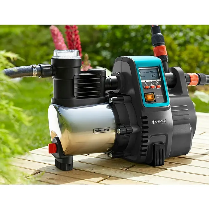 Gardena Bewässerungspumpe Haus- & Gartenautomat 6000/6 LCD inox - 1.300 W, Max. Fördermenge: 6.000 l/h - Nahaufnahme einer Gardena 6000/6E LCD Inox Hauswasserautomat Pumpe auf einer Holzterrasse mit Gartenhintergrund.