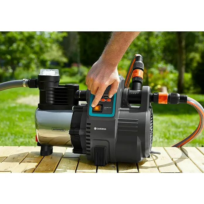 Gardena Bewässerungspumpe Haus- & Gartenautomat 6000/6 LCD inox - 1.300 W, Max. Fördermenge: 6.000 l/h - Nahaufnahme einer Gardena 6000/6E LCD Inox Gartenpumpe, die auf einer Holzoberfläche steht und von einer Hand bedient wird.