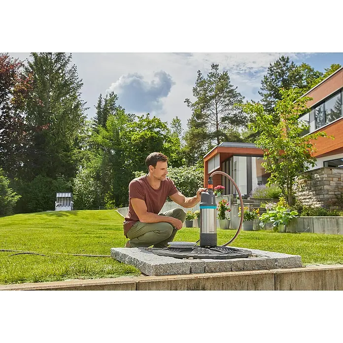 Gardena Tauchpumpe 5900/4 INOX Automatic - 900 W, Max. Fördermenge: 5.900 l/h - Ein Mann hockt neben einer Gardena-Tauchdruckpumpe, die auf einer Steinplattform steht, im Garten, umgeben von üppigem Grün und einem modernen Haus.