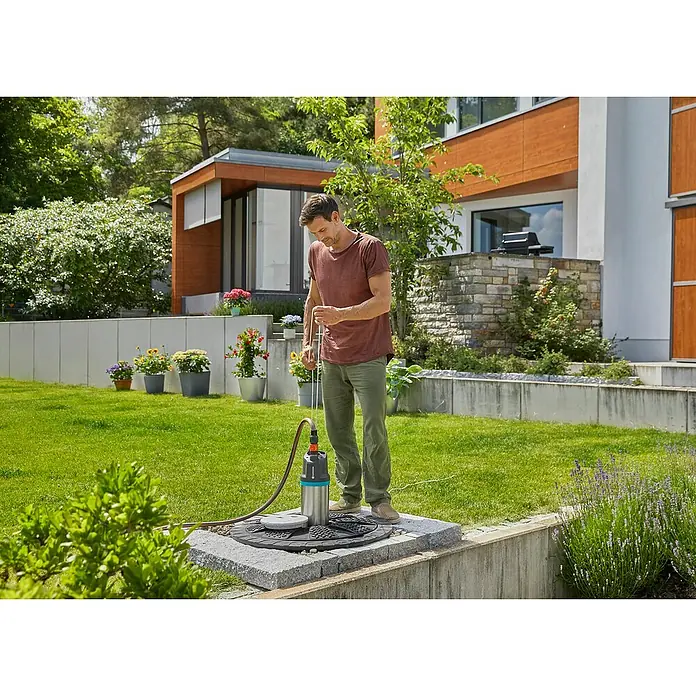 Gardena Tauchpumpe 5900/4 INOX Automatic - 900 W, Max. Fördermenge: 5.900 l/h - Mann bedient eine GARDENA Schmutzwasser-Tauchpumpe auf einer gepflasterten Fläche im Garten, umgeben von Grünflächen und einem modernen Haus.