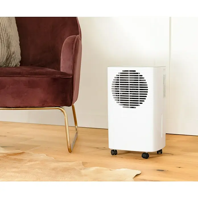 Voltomat Elektro-Luftentfeuchter - 205 W, Entfeuchtungsleistung: 10 l/Tag, L x B x H: 20,5 x 27 x 40,8 cm, Weiß - Weißer Luftentfeuchter mit runder Lüfterabdeckung neben einem burgunderroten Samtsessel auf Holzboden.
