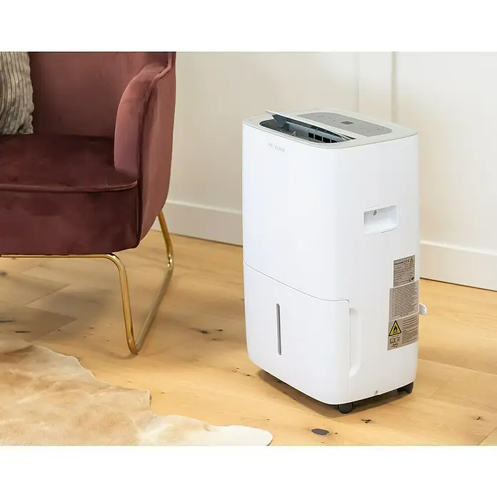 Voltomat Elektro-Luftentfeuchter mit Schuhtrockner - 520 W, Entfeuchtungsleistung: 30 l/Tag, L x B x H: 31,5 x 39,5 x 53,5 cm, Weiß, Grau - Weißer ProKlima Luftentfeuchter steht neben einem burgunderfarbenen Samtsessel auf einem Holzboden.