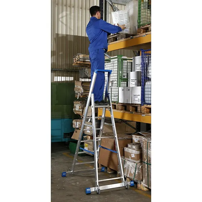 Krause Stabilo Stehleiter Fahrbar mit Traverse - Anzahl Tritte: 6 Stk., Arbeitshöhe: 3,4 m, Aluminium - Eine Person in blauer Arbeitskleidung steht auf einer fahrbaren Leiter und hantiert mit weißen Behältern in einem Lagerregal.