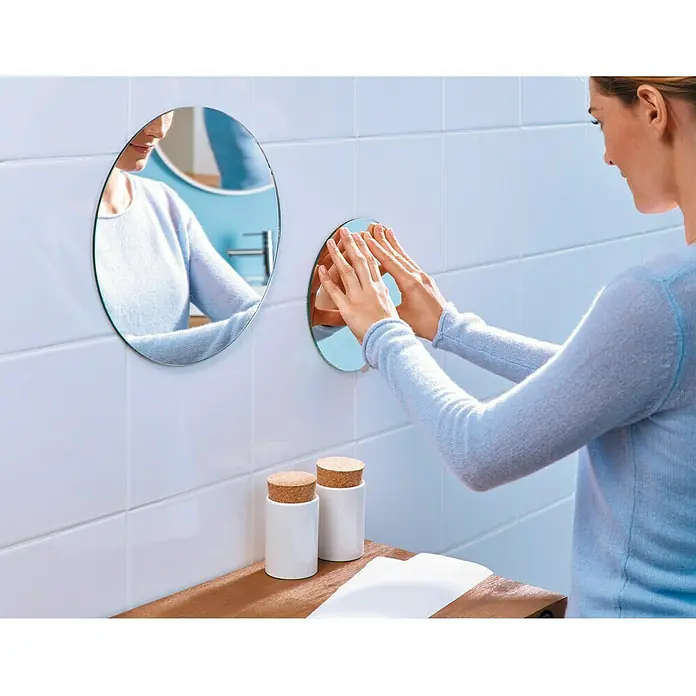 Tesa Powerbond Cinta de doble cara Resistente humedad - 1,9 cm x 5 m, Blanco - Primer plano de una mujer que coloca un espejo de pared redondo en el baño, junto al cual hay recipientes blancos con tapones de corcho.