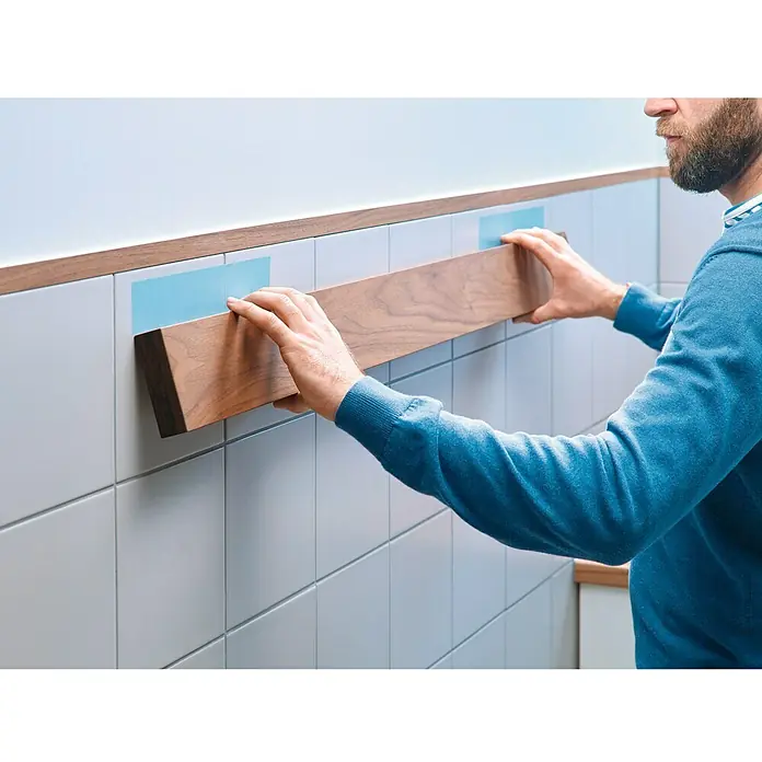 Tesa Powerbond Cinta de doble cara Azulejos/Metal - L x An: 5 m x 1,9 cm, Blanco - Primer plano de una persona fijando una tabla de madera a una pared de azulejos marcada con indicadores azules, posiblemente como soporte o adorno.