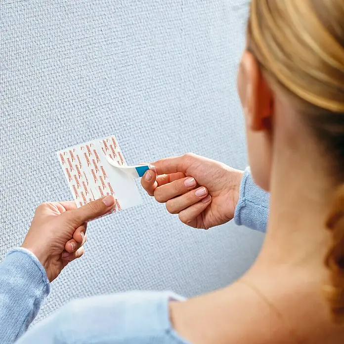 Tesa Clavo adhesivo - Apto para: Paredes pintadas, Carga soportada: 1 kg, 2 ud., Blanco - Una mujer, vista desde atrás, está fijando una tira adhesiva Command a una pared con textura. Ideal para colgar cuadros.
