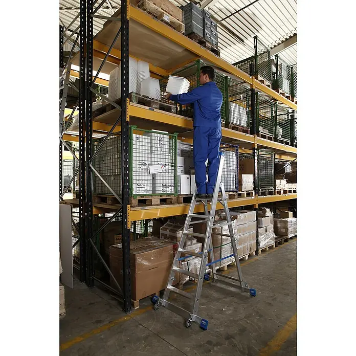 Krause Stabilo Stehleiter Fahrbar mit Traverse - Anzahl Tritte: 6 Stk., Arbeitshöhe: 3,4 m, Aluminium - Ein Mann steht auf einer Aluleiter vor Lagerregalen und sortiert weiße Behälter. Die Leiter ist mit Rollen für mehr Mobilität ausgestattet.