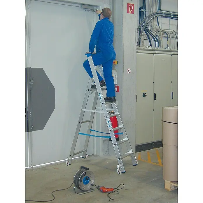 Krause Stabilo Stehleiter Fahrbar - Anzahl Tritte: 6 Stk., Arbeitshöhe: 3,4 m, Aluminium - Eine Person in blauer Arbeitskleidung steht auf einer Krause-Leiter und arbeitet an einer Tür. Die Leiter verfügt über Rollen und Sicherheitsmerkmale.