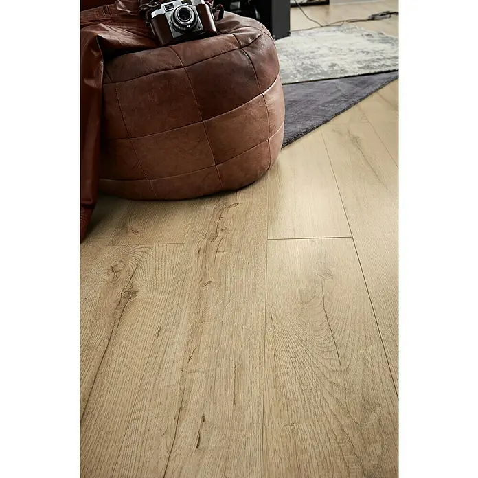 LOGOCLIC Laminado Roble Selva - 1.380 x 193 x 10 mm, Roble Selva, AC5 - Primer plano de un puf de cuero marrón con una cámara vintage sobre un suelo de madera clara, creando un ambiente acogedor.