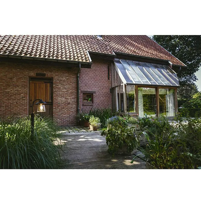 Lutec Lámpara de pie para exterior Cate - 40 W, Color del marco: Antracita, 26,5 x 17,6 x 94,5 cm, Sin bombillas - Una lámpara de exterior atmosférica de metal oscuro frente a un edificio de ladrillo con plantas y un jardín de invierno.