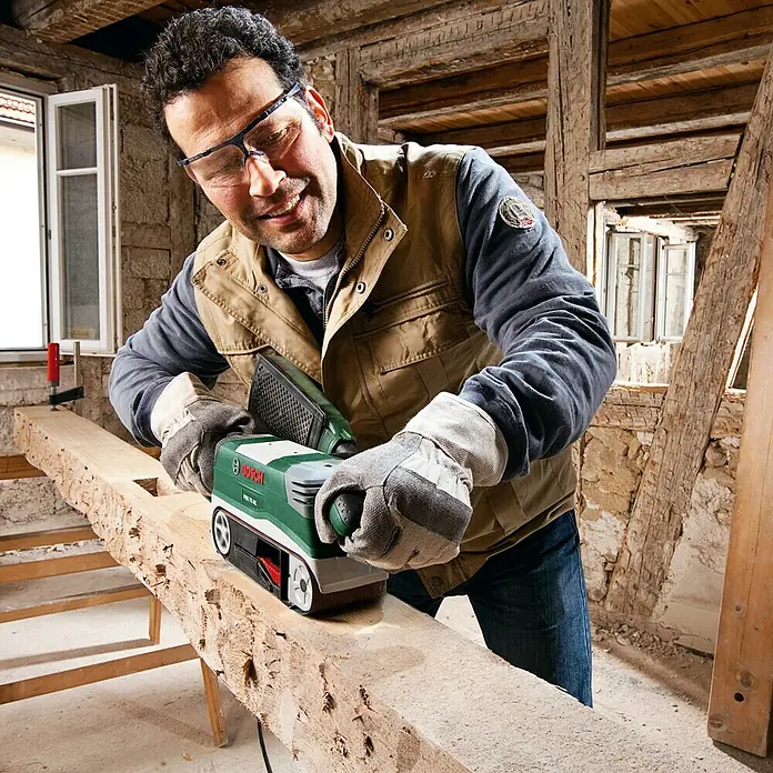 Bosch Elektro-Bandschleifmaschine PBS 75 AE Set - 750 W - Ein Mann verwendet einen grünen Bosch Prio 75 AE Bandschleifer, um ein Holzbrett zu schleifen, wobei er Schutzbrille und Handschuhe trägt.