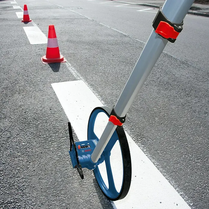 Bosch Professional Messrad GWM 32 - Messbereich: Max. 9999,99 m, Messgenauigkeit: 10 cm/100 m - Nahaufnahme eines Bosch-Messrads auf einer Straße, das zum genauen Messen von Distanzen verwendet wird. Verkehrskegel sind im Hintergrund.
