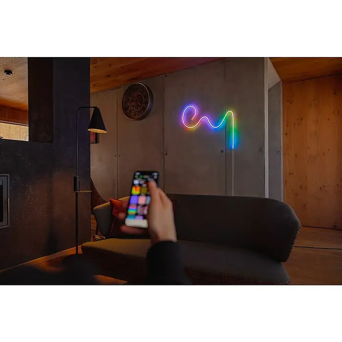 Twinkly Tira de luces LED Flex - Largo: 2 m, Color de luz: RGB, 15 W - Mano con smartphone al frente, al fondo luz de neón de colores en la pared, creando un ambiente acogedor. El foco está en la iluminación inteligente.