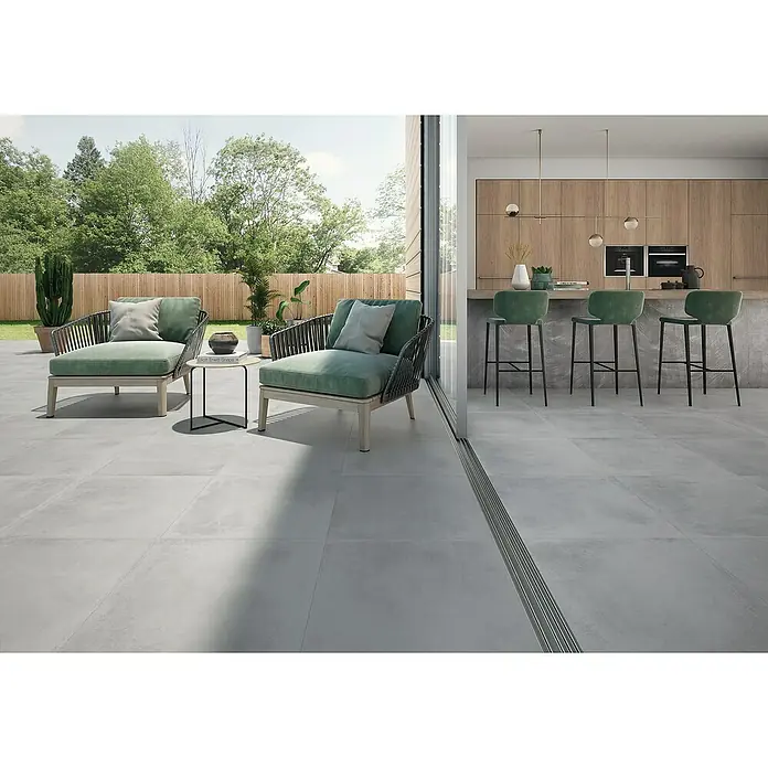 BHS Showroom Pavimento porcelánico Madox In&Out - 60,5 x 60,5 cm, Gris, Mate - Interior lujoso con baldosas grises, cómodos sillones y una cocina moderna con armarios de madera y taburetes de bar verdes.