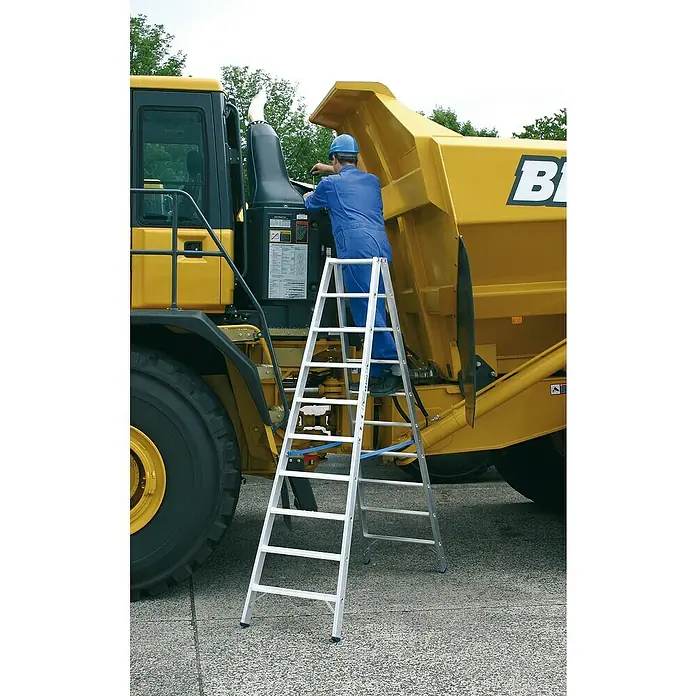 Krause Stabilo Doppelleiter - Anzahl Tritte: 20 Stk., Arbeitshöhe: 3,9 m, Aluminium - Ein Arbeiter steht auf einer silbernen Leiter und repariert einen gelben Bagger. Die Leiter steht stabil auf dem Boden.