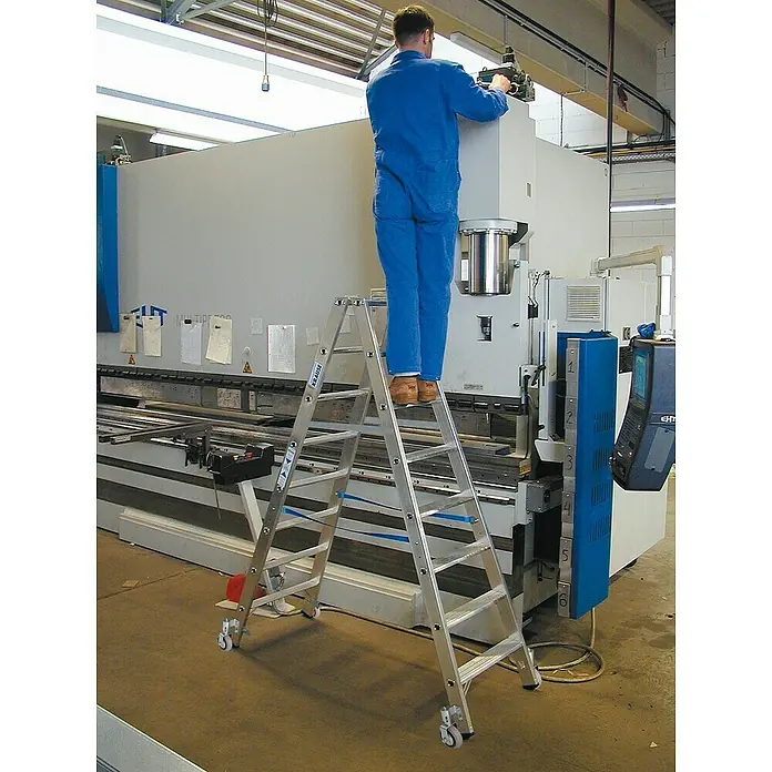 Krause Stabilo Doppelleiter Fahrbar - Anzahl Tritte: 16 Stk., Arbeitshöhe: 3,4 m, Aluminium - Eine rollbare Krause Leiter vor einer großen Industriemaschine, ein Mann in Arbeitskleidung steht darauf und arbeitet an der Maschine.