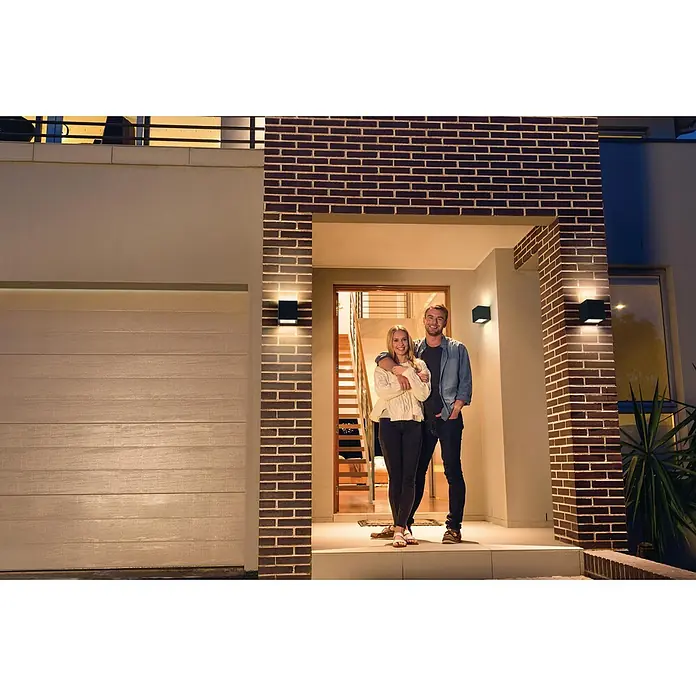 Ledvance Smart+ WiFi LED-Außenwandleuchte BRICKFarbe Gehäuse: Dunkelgrau, IP44 - Fassadenleuchten beleuchten ein modernes Haus, während ein Paar auf der Veranda steht. Die quadratischen Leuchten akzentuieren das Mauerwerk stilvoll.