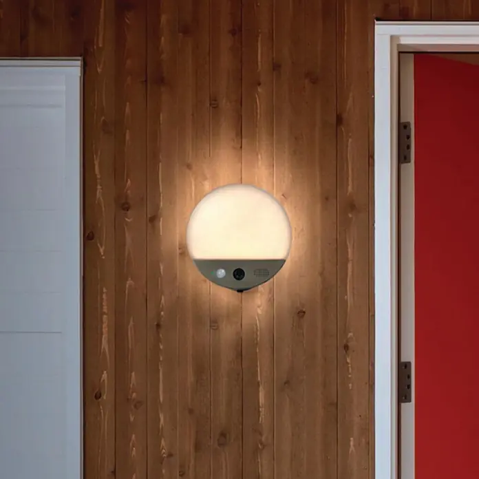 Osram LED-Außenwandleuchte105 x 180 x 105 mm, Kamera, Silber - Diese Nahaufnahme zeigt eine Wandleuchte mit Bewegungssensor, die an einer Holzwand neben einer Tür montiert ist und für Beleuchtung sorgt.