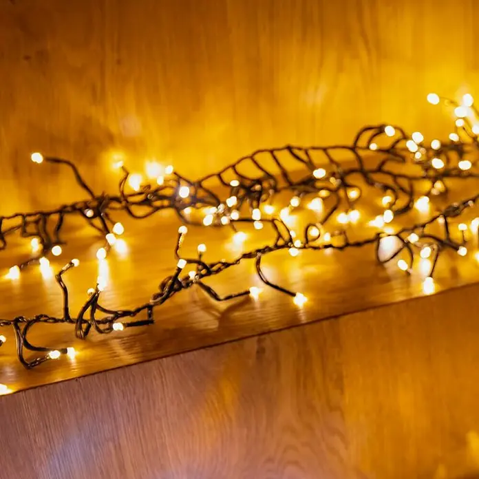 Twinkly Cadena de luces LED Cluster - Para exterior, Largo: 6 m, 400 ud., Funcionamiento en red, Color de luz: Ámbar - Guirnalda de luces decorativa con LEDs blanco cálido sobre una estantería de madera, creando un ambiente acogedor.