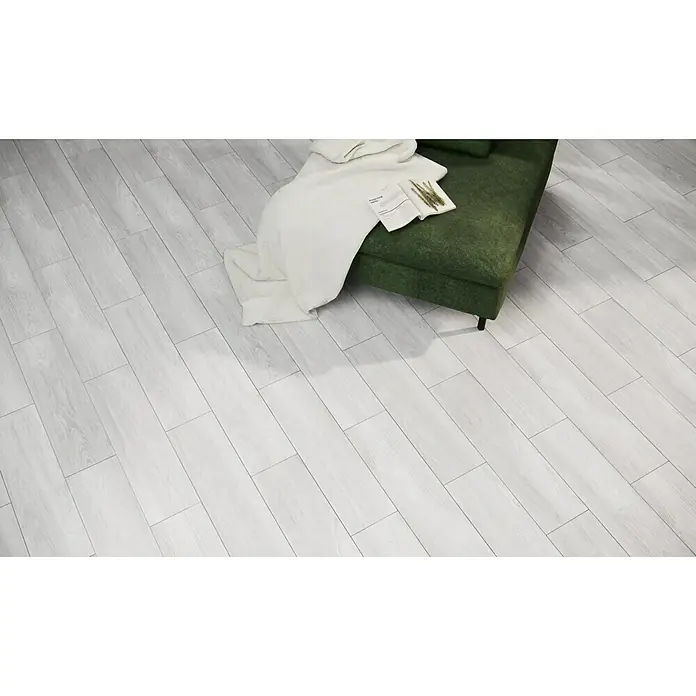 Pavimento porcelánico Alcázar - 20 x 75 cm, Pearl, Mate - Un salón luminoso y moderno con un sofá verde y un libro sobre una manta blanca. El suelo es de tablones de madera de color claro.