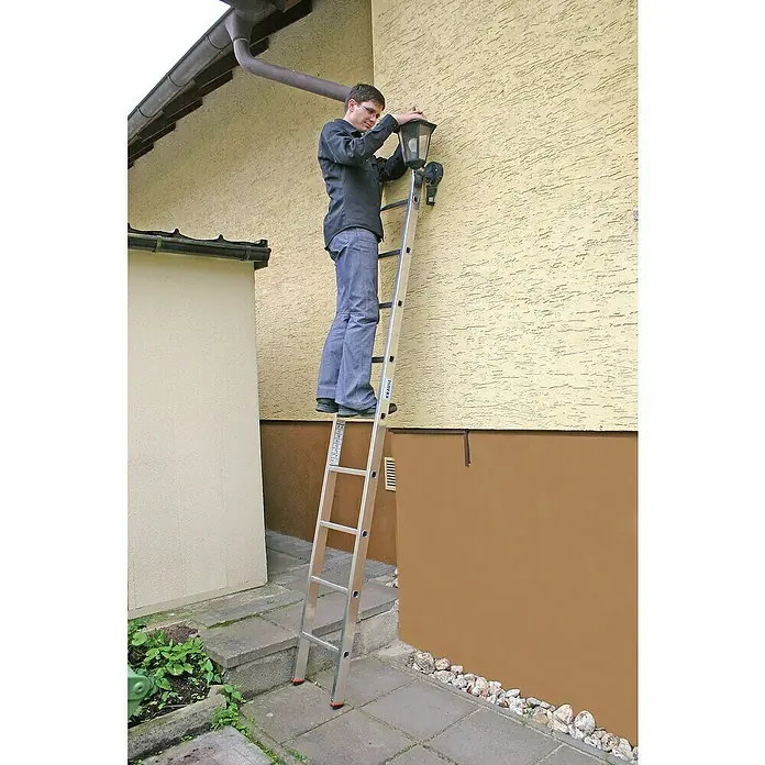 Krause Monto Anlegeleiter Sibilo - Anzahl Tritte: 9 Stk., Arbeitshöhe: 3,6 m, Belastbarkeit: 150 kg, Aluminium - Mann auf einer Krause Leiter, der eine Lampe an einer beigen Wand befestigt. Die Leiter steht sicher auf Steinplatten und bietet Halt für Reparaturen.