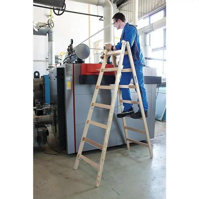 Krause Stabilo Escalera doble - Número de peldaños: 14 ud., Altura de trabajo: 3,25 m, Carga soportada: 150 kg, Madera - Hombre en escalera de madera frente a una caldera. Entorno laboral seguro, equipamiento fiable.