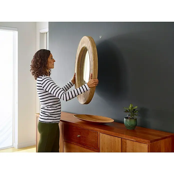 3M Claw Gancho para colgar cuadros - 2 ud., Capacidad de carga: 11 kg - Mujer que cuelga un espejo redondo con marco de madera en una pared gris sobre un aparador de madera.