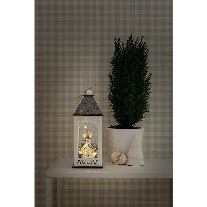 Konstsmide LED-Weihnachtsdekoleuchte - Weiß, Batteriebetrieben, LED - Festliche Szene mit einer beleuchteten Laterne, die eine Weihnachtsszene zeigt, neben einer Zimmerpflanze und dekorativen Elementen auf einem Tisch.