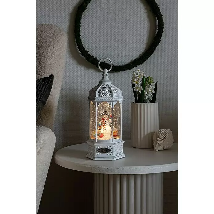 Konstsmide LED-Schneelaterne - Innen, Weiß, 13 x 11 x 29 cm, Schneemann, Batteriebetrieben - Weiße Laternen-Schneekugel mit beleuchtetem Schneemann auf einem weißen Beistelltisch.