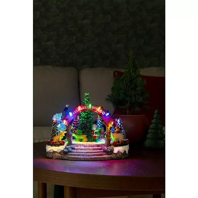 Konstsmide LED-Szenerie Weihnachtszoo - L x B x H: 28 x 22,5 x 20 cm, Netzbetrieben, LED - Ein farbenfrohes, beleuchtetes Weihnachtsdorf mit einem 'WELCOME TO THE ZOO'-Bogen und animierten Figuren, auf einem Holztisch.