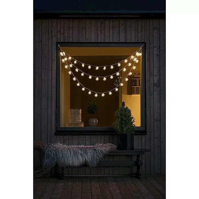 Konstsmide LED-Lichtervorhang - Außen, 1,25 m, Warmweiß, 44 Stk., Netzbetrieben - Warmweiße, schneeflockenförmige Lichterkette, die über einem dunklen Fenster hängt und den Außenbereich beleuchtet.