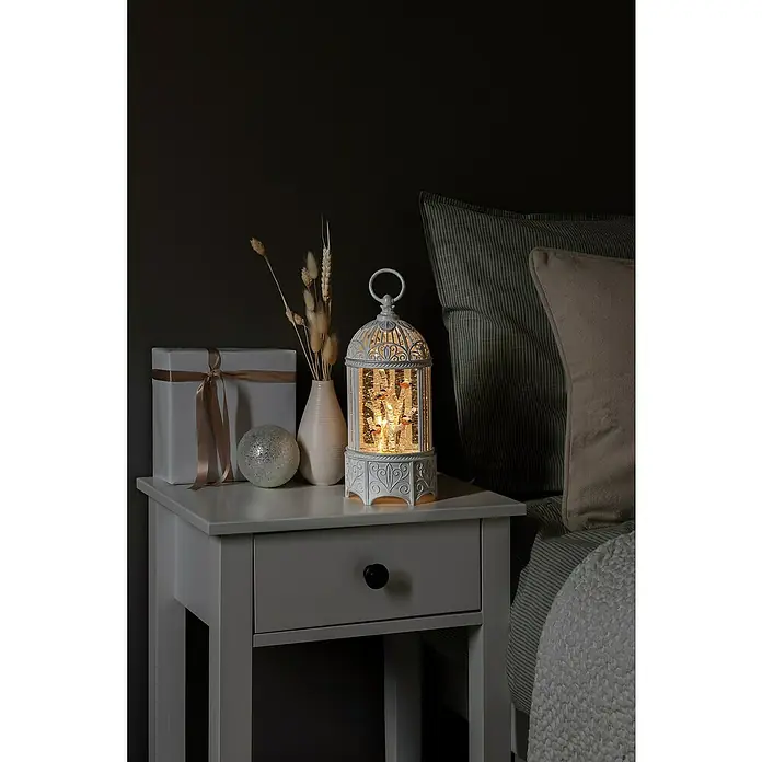 Konstsmide LED-Schneelaterne Gipfel im Baum - Innen, Weiß, 12 x 12 x 25,5 cm, Batteriebetrieben - Dekorative Szene mit einer beleuchteten, weißen Laterne mit Wintermotiv auf einem Nachttisch neben einem Bett. Geschenk, Vase und Kugel runden das Bild ab.