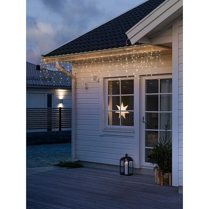 Konstsmide LED-Lichtervorhang Eisregen - Außen, Warmweiß, 80 Stk., Batteriebetrieben - Festliche Außenansicht eines Hauses mit funkelnden Eiszapfenlichtern und einem leuchtenden Stern im Fenster.