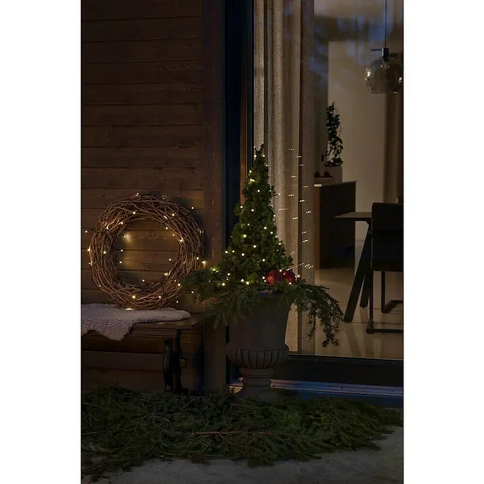 Konstsmide LED-Lichterkette - Außen, Lichtfarbe: Warmweiß, Anzahl Leuchtmittel: 120 Stk., Batteriebetrieben - Stimmungsvolle Weihnachtsdekoration mit leuchtendem Kranz und Mini-Weihnachtsbaum auf einer dunklen Holzveranda.